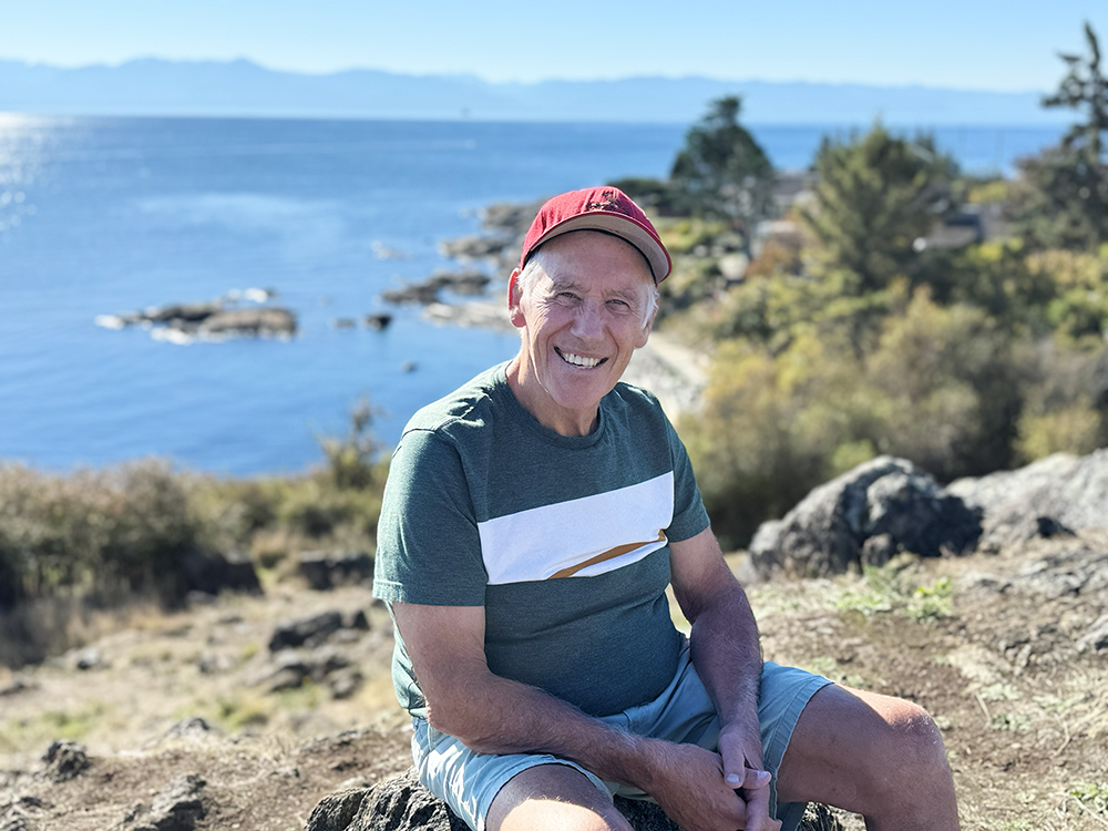 Peter sitting on a rock with the ocean and mountains behind him. 